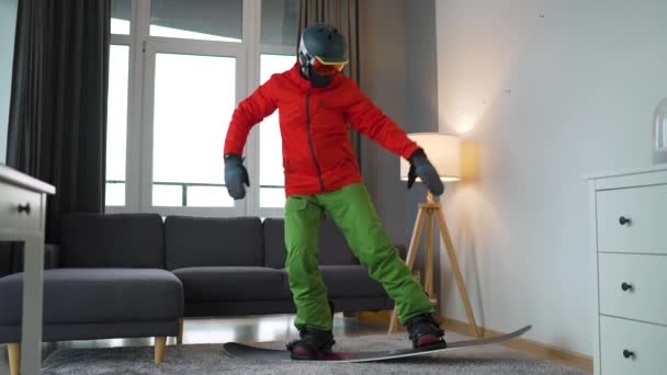 Vídeo divertido. Hombre vestido de snowboarder representa el snowboard en una alfombra en una habitación acogedora. Esperando un invierno nevado. Movimiento lento — Vídeo de stock