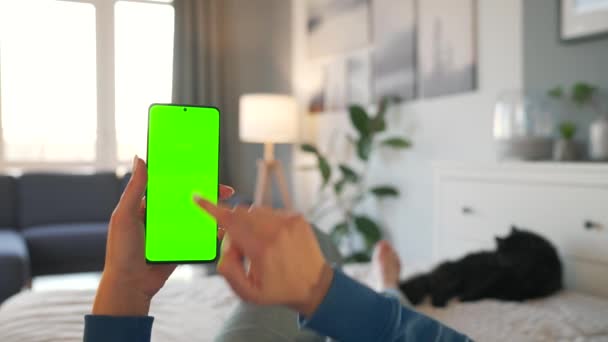 Žena doma leží na posteli s černou kočkou a používá smartphone se zelenou maketou obrazovky ve vertikálním režimu. Dívka procházející internet, sledující obsah, videa. POV. — Stock video