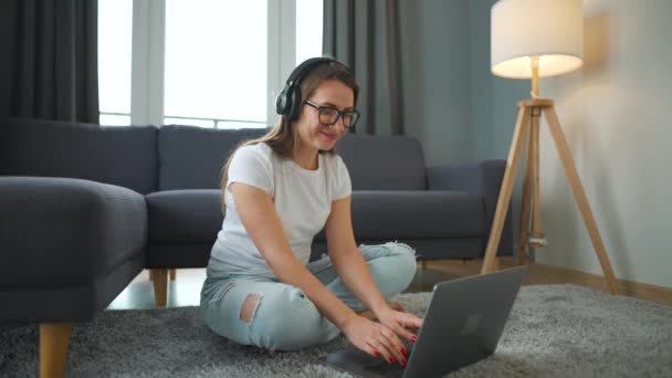 Casualmente mujer vestida con auriculares está sentado en la alfombra con el ordenador portátil y trabajando en una habitación acogedora. Trabajo remoto fuera de la oficina. — Vídeos de Stock