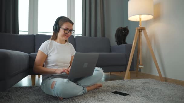 Alkalmanként öltözött nő fülhallgatóval ül a szőnyegen laptoppal, és távolról dolgozik a hangulatos szobában. Mögötte a kanapén fekszik egy fekete macska. — Stock videók