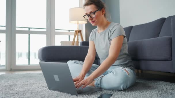 Lässig gekleidete Frau sitzt auf Teppich mit Laptop und Smartphone und arbeitet in gemütlichem Raum. Fernarbeit außerhalb des Büros. — Stockvideo