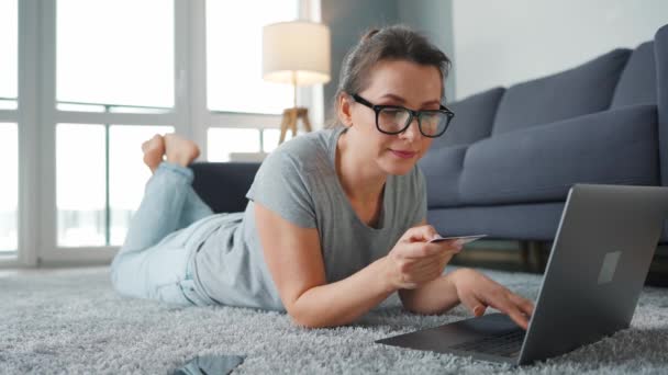 Donna con gli occhiali è sdraiata sul pavimento e fa un acquisto online con carta di credito e laptop. Shopping online, tecnologia lifestyle — Video Stock