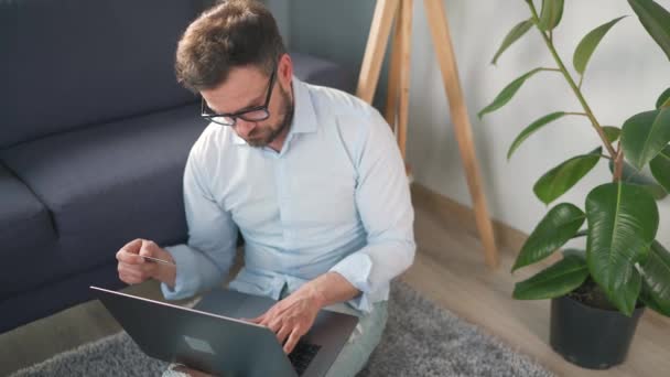 Hombre con gafas sentado en el suelo y hace una compra en línea con una tarjeta de crédito y un ordenador portátil. Compras en línea, tecnología de estilo de vida — Vídeos de Stock