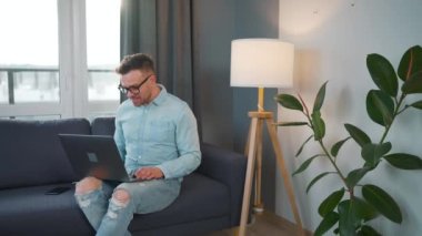 Kanepede oturan gözlüklü sıradan giyinmiş bir adam ve modern bir dairede evden bir video görüşmesi yapmak için laptop kullanıyor.