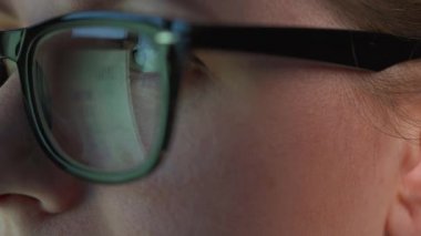 Gözlüklü kadın monitöre bakıyor ve çizelgeler ve analizlerle çalışıyor. Monitör ekranı gözlüklere yansıyor. Gece çalış. Aşırı yakın çekim