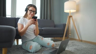 Kulaklık takan sıradan giyinmiş bir kadın dizüstü bilgisayarı ve akıllı telefonuyla halıda oturuyor ve rahat bir odada çalışıyor. Ofis dışında uzaktan çalışma.