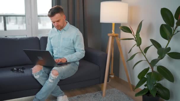 Hombre con gafas sentado en el sofá con ordenador portátil y trabajando en una habitación acogedora. Trabajo remoto fuera de la oficina. — Vídeos de Stock