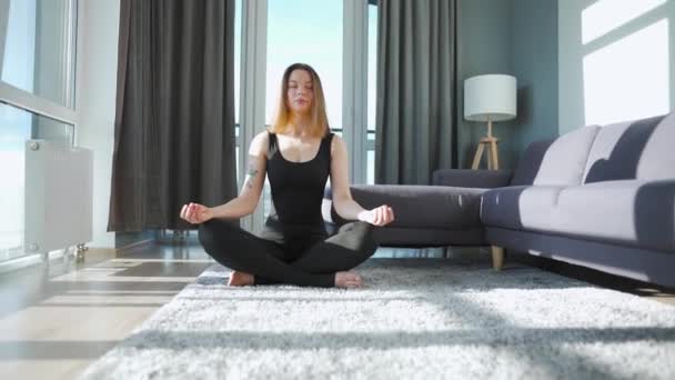 Fiatal fehér nő fekete kezeslábasban lótuszpozícióban ül és meditál, jógázik otthon. Lassú mozgás. — Stock videók