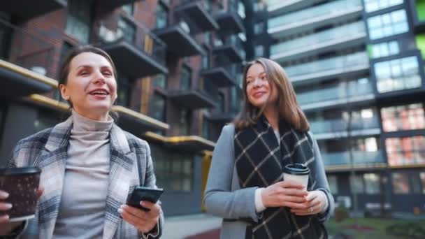 Zwei fröhliche Frauen spazieren mit Kaffee zum Mitnehmen durch das Geschäftsviertel und unterhalten sich interessiert untereinander. Zeitlupe — Stockvideo