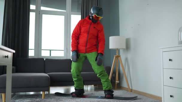 Hezké video. Muž oblečený jako snowboardista zobrazuje snowboarding na koberci v útulné místnosti. Čekání na sněhovou zimu. — Stock video