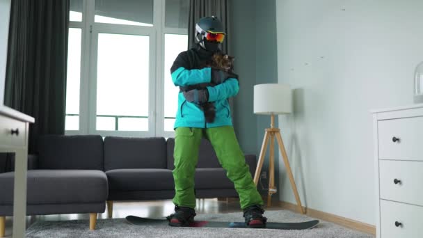 Vídeo divertido. Hombre vestido de snowboarder monta una tabla de snowboard en una alfombra en una habitación acogedora. Tiene un gato esponjoso en sus brazos. Esperando un invierno nevado. Movimiento lento — Vídeo de stock