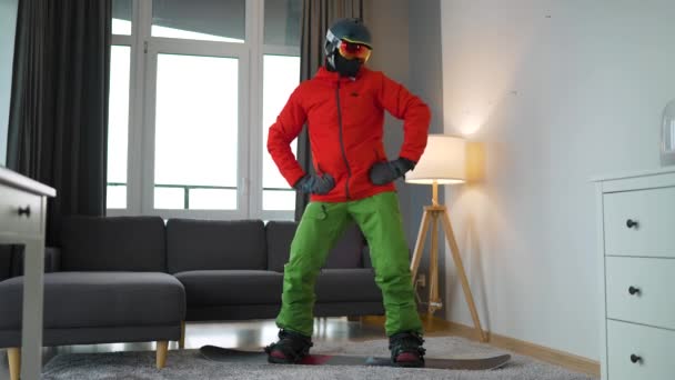 Vídeo divertido. Hombre vestido como un snowboarder felizmente bailando y divirtiéndose mientras está de pie sobre una tabla de snowboard en una alfombra en una habitación acogedora. Esperando un invierno nevado. Movimiento lento — Vídeo de stock