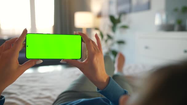 Mujer en casa acostada en una cama y usando un smartphone con pantalla verde simulada en modo horizontal. Chica navegando por Internet, viendo contenido, videos. — Vídeo de stock