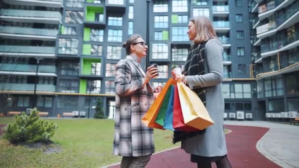 Δύο ευτυχισμένες γυναίκες στέκονται με τσάντες για ψώνια και παίρνουν καφέ μετά από επιτυχημένα ψώνια και μιλάνε με ενδιαφέρον μεταξύ τους.. — Αρχείο Βίντεο