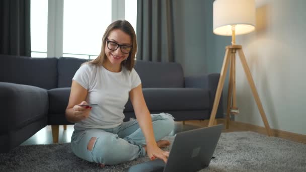 Donna con gli occhiali si siede sul pavimento e fa un acquisto online con carta di credito e laptop. Shopping online, tecnologia lifestyle — Video Stock