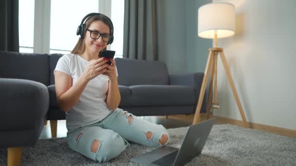 Casualmente mujer vestida con auriculares está sentado en la alfombra con el ordenador portátil y el teléfono inteligente y trabajando en una habitación acogedora. Trabajo remoto fuera de la oficina. — Vídeos de Stock