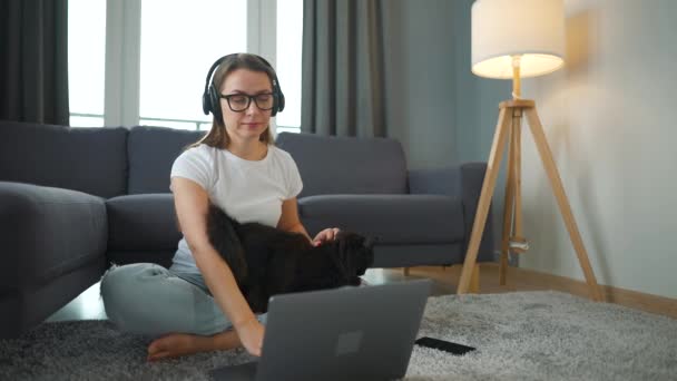 Mujer vestida casualmente se sienta en una alfombra con un ordenador portátil, se sujeta las rodillas y acaricia a un gato esponjoso y trabaja en una habitación acogedora. Trabajo remoto fuera de la oficina. — Vídeos de Stock
