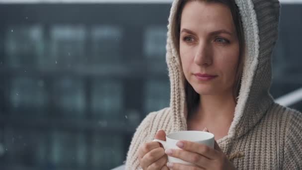 Kaukasische Frau hält sich bei Schneefall mit heißem Kaffee oder Tee auf dem Balkon auf. Sie blickt auf die Schneeflocken und atmet die frostige frische Luft ein — Stockvideo
