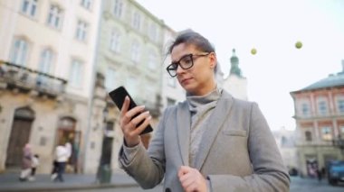 Gözlüklü bir kadın eski bir caddede yürürken ve akıllı telefon kullanırken.
