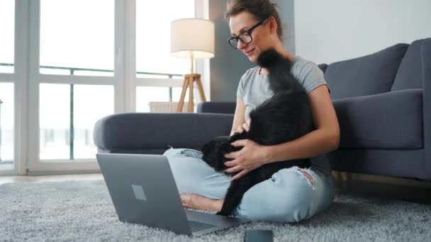 Випадково одягнена жінка сидить на килимі з ноутбуком, тримається на колінах і гладить пухнастого кота і працює в затишній кімнаті. Віддалена робота за межами офісу . — стокове відео