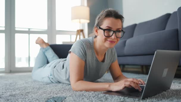 Gözlüklü kadın yerde yatıyor ve dizüstü bilgisayar üzerinde çalışıyor. Uzak çalışma kavramı. — Stok video