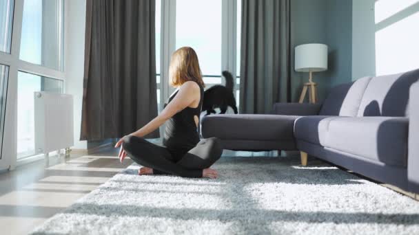 Jonge blanke vrouw in zwarte jumpsuit doet yoga thuis om flexibiliteit en evenwicht te ontwikkelen. Zwarte kat zit op de achtergrond. Langzame beweging — Stockvideo