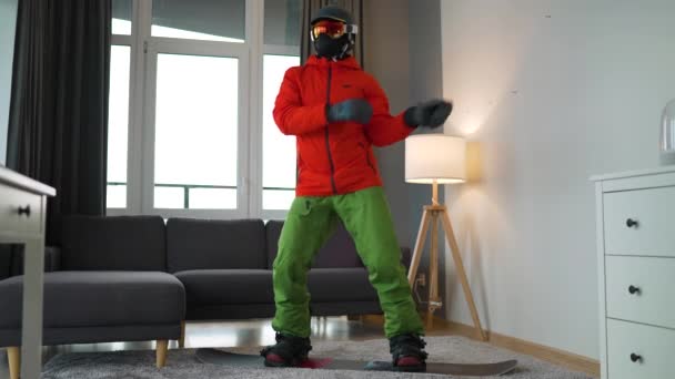 Vídeo engraçado. Homem vestido como um snowboarder feliz dançando e se divertindo enquanto está de pé em um snowboard em um tapete em um quarto acolhedor. À espera de um Inverno nevado. Movimento lento — Vídeo de Stock