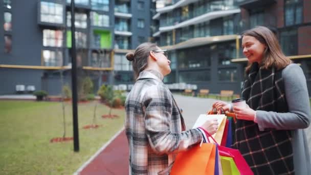 Dos mujeres felices se paran con bolsas de compras y café para llevar después de una compra exitosa y hablan con interés entre ellas. Movimiento lento — Vídeos de Stock