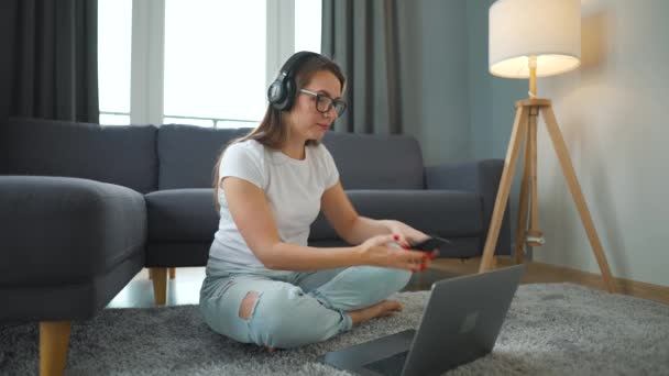 Donna vestita con disinvoltura con le cuffie è seduto su tappeto con computer portatile e di lavoro in camera accogliente. Lavoro a distanza fuori dall'ufficio. — Video Stock