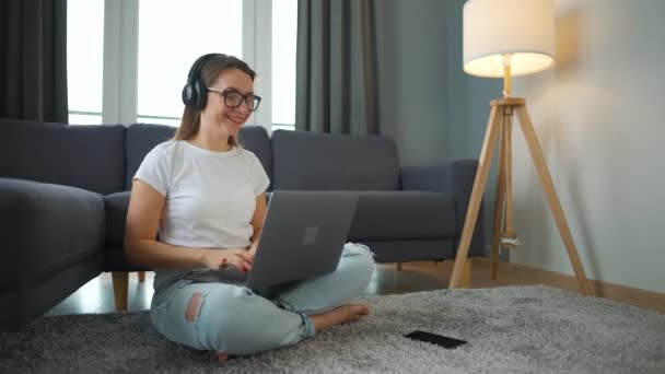 Alkalmanként öltözött nő fülhallgatóval ül a szőnyegen laptoppal, és dolgozik a hangulatos szobában. Távoli munka az irodán kívül. — Stock videók