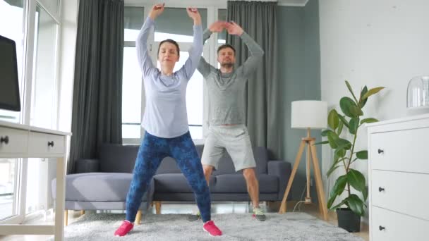 Kaukaskie para robi skoków jacks ćwiczenia w domu w przytulny jasny pokój. — Wideo stockowe