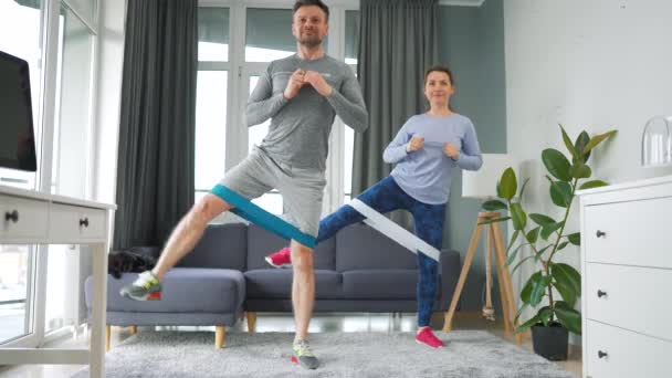 Couple caucasien fait squats et jambes balançoires avec bande élastique à la maison dans une chambre lumineuse confortable. — Video