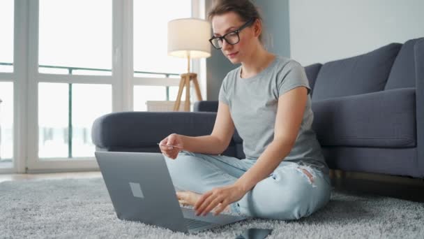Mujer con gafas se sienta en el suelo y hace una compra en línea con una tarjeta de crédito y un ordenador portátil. Compras en línea, tecnología de estilo de vida — Vídeos de Stock