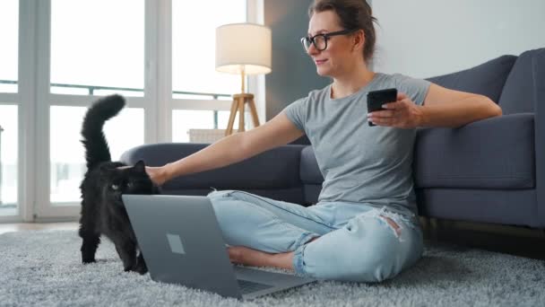 穿着随意的女人坐在地毯上，带着笔记本电脑，跪下来抚摸一只毛绒绒的猫，在一个舒适的房间里工作。办公室外的远程工作. — 图库视频影像
