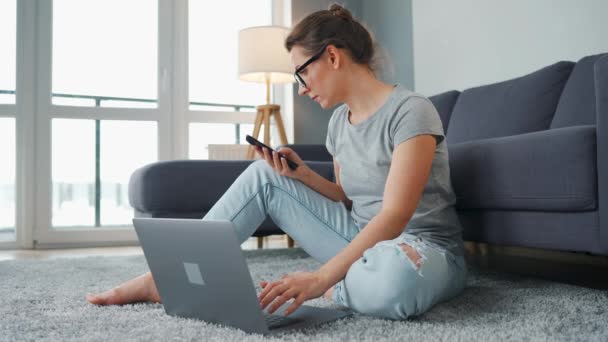 Vrijetijds geklede vrouw zittend op tapijt met laptop en smartphone en werkend in gezellige kamer. Werk op afstand buiten het kantoor. — Stockvideo