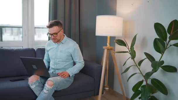 Casual habillé homme avec des lunettes assis sur le canapé et en utilisant un ordinateur portable pour faire un appel vidéo de la maison dans un appartement moderne — Video