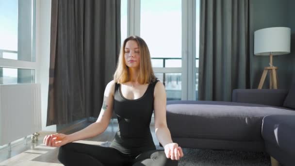 Mujer joven caucásica en mono negro sentada en posición de loto y meditando, yoga en casa. Movimiento lento — Vídeos de Stock