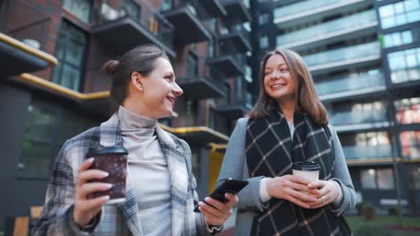 Dua wanita bahagia berjalan dengan membawa kopi dan berbicara dengan minat di antara mereka sendiri di distrik bisnis. — Stok Video