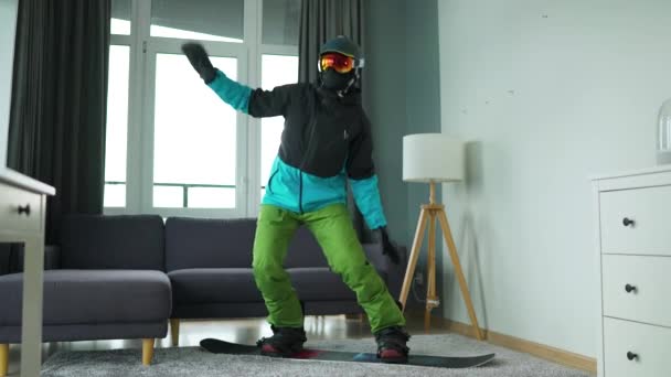 Un vídeo divertido. Hombre vestido de snowboarder representa el snowboard en una alfombra en una habitación acogedora. Esperando un invierno nevado. Movimiento lento — Vídeo de stock