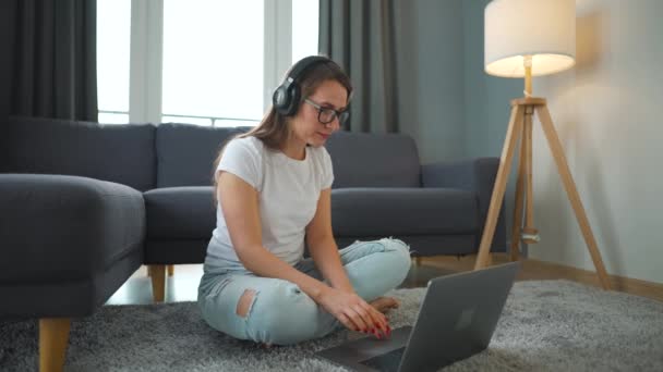 Casually ντυμένη γυναίκα με ακουστικά κάθεται στο χαλί με φορητό υπολογιστή και εργάζονται σε άνετο χώρο. Απομακρυσμένη εργασία έξω από το γραφείο. — Αρχείο Βίντεο
