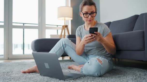 Lässig gekleidete Frau sitzt auf Teppich mit Laptop und Smartphone und arbeitet in gemütlichem Raum. Fernarbeit außerhalb des Büros. — Stockvideo