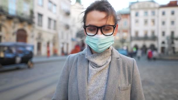 Pandemische bescherming van het Covid-19 coronavirus. Portret van een vrouw in een jas, bril en een beschermend medisch masker. Ze staat midden op het plein. Langzame beweging — Stockvideo