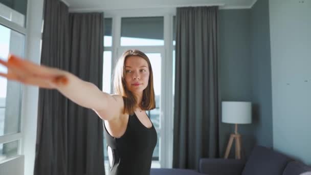 Jonge blanke vrouw in zwarte jumpsuit doet yoga virabhadrasana thuis om flexibiliteit en evenwicht te ontwikkelen. Langzame beweging — Stockvideo