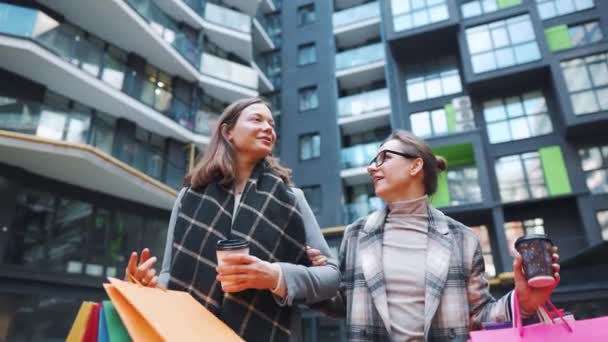 Due donne felici camminano con borse della spesa e caffè da asporto dopo uno shopping di successo e parlano con interesse tra di loro. Rallentatore — Video Stock