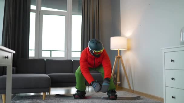 Un vídeo divertido. Hombre vestido de snowboarder representa el snowboard en una alfombra en una habitación acogedora. Esperando un invierno nevado. Movimiento lento — Vídeos de Stock