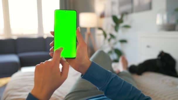 自宅の女性は黒い猫とベッドの上に横になり、垂直モードで緑色のモックアップ画面を持つスマートフォンを使用しています。女の子はインターネットを閲覧し、コンテンツ、ビデオを見る。POV. — ストック動画