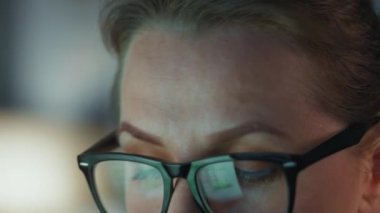 Gözlüklü kadın monitöre bakıyor ve çizelgeler ve analizlerle çalışıyor. Monitör ekranı gözlüklere yansıyor. Gece çalış. Aşırı yakın çekim