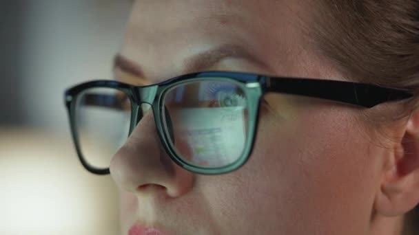 안경을 쓴 여성 이 모니터를 보고 차트와 분석 자료를 가지고 일하고 있습니다. 안경에는 모니터가 반사되어 있다. 밤에는 일하고. 극도로 근접하다 — 비디오