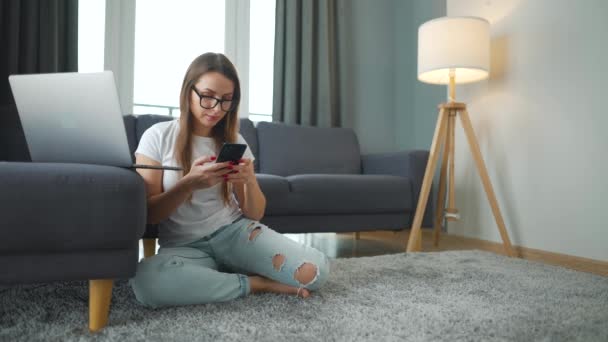 Wanita berpakaian santai duduk di karpet dengan laptop dan smartphone dan bekerja di kamar yang nyaman. Pekerjaan jarak jauh di luar kantor. — Stok Video
