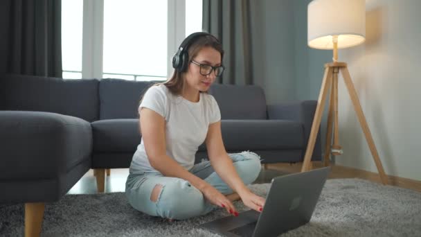 Casually klädd kvinna med hörlurar sitter på matta med laptop och arbetar i mysiga rum. Fjärrarbete utanför kontoret. — Stockvideo